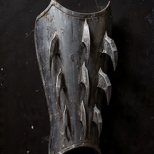 May include: A weathered, dark metal arm guard with a series of sharp, claw-like protrusions. The surface shows signs of wear, suggesting a medieval or fantasy theme. The design is curved to fit the arm.