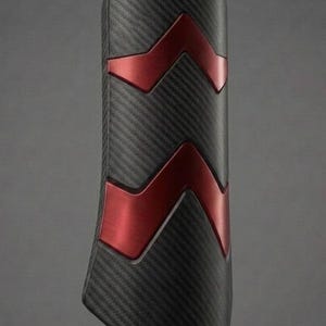 May include: A protective shin guard featuring a black carbon fiber texture finish with two prominent, raised, metallic red chevron-shaped accents positioned vertically along the center of the curved, rectangular protective shell.