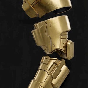 May include: A set of metallic gold armour components including a rounded shoulder piece, an upper arm guard, a forearm gauntlet with segmented detailing, and a separate hand plate, all featuring a brushed finish and geometric panel lines.