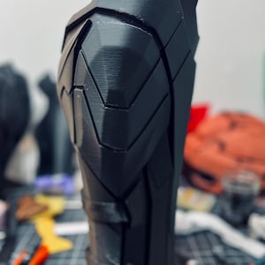 May include: A black, angular, 3D-printed armour piece, possibly a leg or arm guard. The design features geometric panels and a textured surface. The piece is standing upright, with a blurred background of tools and other objects.