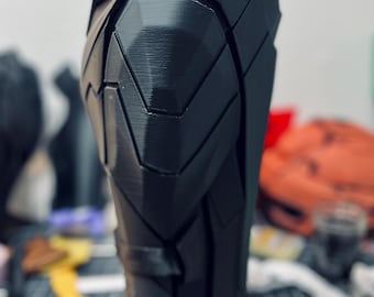 May include: A black, angular, 3D-printed armor piece, possibly a leg or arm guard. The design features geometric panels and a textured surface. The piece is standing upright, with a blurred background of tools and other objects.