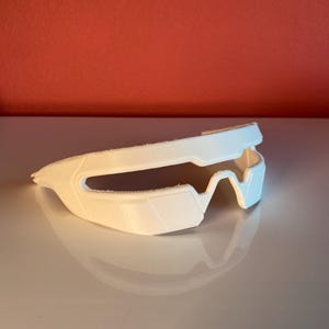 May include: A white, single-piece frame structure with a wrap-around design, featuring a wide, horizontal opening across the front and a bridge section for the nose. The object has a matte finish and a rigid, angular construction with visible texture.
