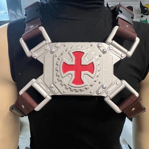 May include: A silver and red chest plate with a cross design, secured by brown straps and buckles. The plate has a gear-like border and is worn over a black top. This item is likely a costume piece or cosplay accessory.