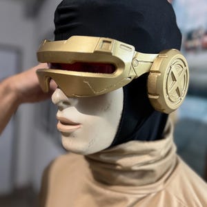 May include: A gold-colored visor with a red lens and circular side pieces featuring an X-shaped emblem. The visor is worn over a black fabric hood on a mannequin head, which is dressed in a tan high-neck garment. The design is rigid and metallic.