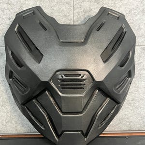 以下が含まれることがあります： マットなチャコールグレーの胸当てアーマーパーツ。構造的で多面的なデザインが特徴で、埋め込み式の通気口と幾何学的なパネルが施されています。表面にはメタリックな光沢があり、中央に水平なグリルを備えた硬質で成形された構造をしています。
