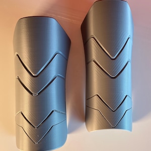 May include: A pair of matte gray shin guards featuring a distinct chevron pattern with three horizontal V-shaped ridges molded into the surface. The guards have a curved, semi-cylindrical shape designed to fit the contours of a lower leg.