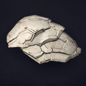 May include: A weathered, off-white shoulder armor piece with a geometric, segmented design. The armor has a distressed finish, suggesting age or battle wear, and is set against a dark background.