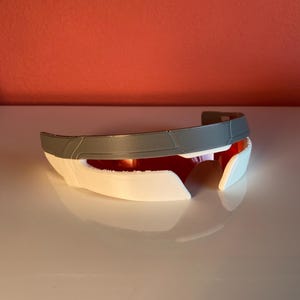 May include: A visor-style eyewear piece featuring a wide, grey upper frame section and a white lower frame section. The lenses are a deep, reflective red color, creating a continuous horizontal band across the front of the accessory.