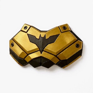 May include: A gold and black chest plate with a bat symbol in the center. The chest plate has a geometric design with four black screws. The chest plate is made of a metallic material.