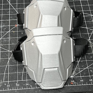 May include: Two silver-colored knee pads with black adjustable straps. The pads have a geometric design with multiple facets and are likely made of a hard, protective material. The pads are designed to protect the knees.