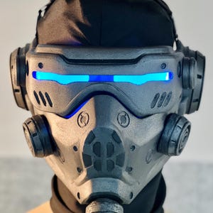 May include: A futuristic, gray and black mask with blue LED lights across the eyes. The mask has a complex design with multiple circular and angular components, suggesting a high-tech or sci-fi theme. A black fabric covers the top of the head.