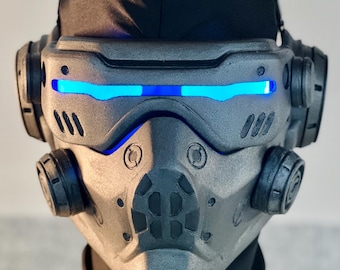 May include: A futuristic, gray and black mask with blue LED lights across the eyes. The mask has a complex design with multiple circular and angular components, suggesting a high-tech or sci-fi theme. A black fabric covers the top of the head.