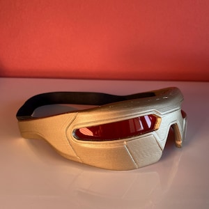 May include: A gold-colored visor with a smooth, molded frame and a single, continuous red lens that spans the front. The visor features a black elastic strap attached to the sides, designed to secure the eyewear around the head for a snug fit.