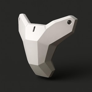 Op de afbeelding: Een witte, geometrische masker met een facetontwerp. Het masker heeft een puntige bovenkant en een gebogen onderkant, met een klein gaatje en een cirkelvormig detail aan de zijkanten. Het masker is gemaakt van een mat materiaal en staat tegen een donkere achtergrond.
