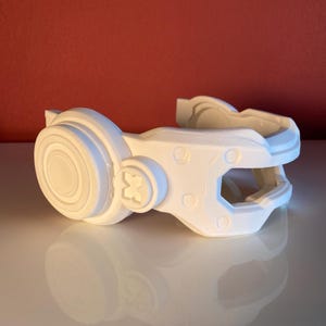 May include: A white 3D printed headset accessory featuring a circular earpiece with a central disc, a small square-shaped emblem on the side, and a structured, angular frame with visible layered textures and geometric cutouts throughout the design.