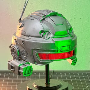 Può includere: Un casco argentato con una luce rossa e accenti verdi. Il casco è su un supporto nero con una base rotonda.