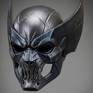 May include: A metallic dark grey full-face helmet featuring a sculpted, aggressive design with prominent pointed ear-like protrusions, deep-set eye sockets, and a lower jaw section detailed with sharp, fang-like teeth and weathered surface textures.