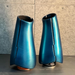 May include: Two teal-colored, metallic-finish objects with a split design. The objects are placed on a gray surface, and the background is a textured, light gray wall. The objects appear to be decorative or functional.