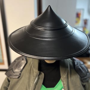 May include: A black conical hat with a wide brim. The hat is worn over a green jacket with black shoulder armor. The hat has a smooth, matte finish and a pointed top. The jacket has a green and black color scheme.