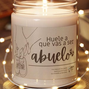 May include: A lit, white candle in a glass jar with a label that reads "Huele a que vas a ser abuelo". The candle sits on a round, wooden coaster. A string of fairy lights and a guitar are in the background.
