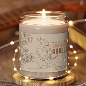 May include: A brown glass candle with a white label that reads "Huele a que vas a ser abuela". The candle is lit and sitting on a wooden coaster. The candle is surrounded by white string lights.