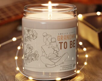 Puede incluir: Una vela de vidrio marrón con una etiqueta blanca que dice "Smells Like a Grandma to Be". La etiqueta presenta un dibujo lineal de una mujer sosteniendo un bebé. La vela está sentada sobre una base de madera.
