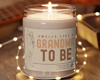 Puede incluir: Una vela de soja marrón con una etiqueta blanca que dice "Smells Like A Grandma To Be". La vela está encendida y se encuentra sobre un posavasos de madera.