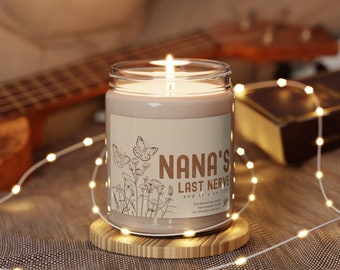 Puede incluir: Una vela marrón con una etiqueta blanca que dice "NANA'S LAST NERVE and it's on fire". La vela está encendida y se encuentra sobre un posavasos de madera. La vela está rodeada de luces blancas.