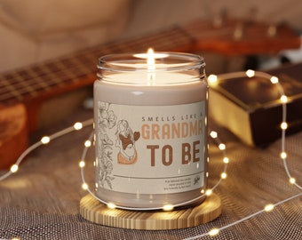Puede incluir: Una vela de soja marrón con una etiqueta que dice "Smells like a Grandma to Be". La etiqueta presenta un dibujo lineal de una mujer embarazada. La vela está sentada sobre un posavasos de madera.