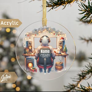 May include: A round, clear acrylic Christmas ornament featuring a cartoon illustration of a person wearing a hooded sweatshirt and headphones, sitting in front of a computer. The ornament has a festive background with lights and a Christmas tree. The text "Name (YEAR) Optional" is included on the ornament.