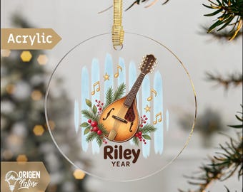 May include: A round clear acrylic Christmas ornament featuring a mandolin illustration, green pine branches, red berries, and gold musical notes. The text "Riley" and "YEAR" is printed in brown. It hangs from a gold ribbon.