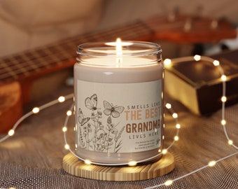 Puede incluir: Una vela marrón con una etiqueta blanca que dice "Smells Like The Best Grandma Lives Here". La etiqueta tiene un diseño floral con mariposas. La vela está sentada sobre un posavasos de madera.