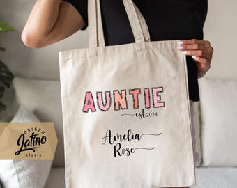May include: A white canvas tote bag with the text "AUNTIE est. 2024 Amelia Rose" printed in pink and black. The text is in a stylized font.