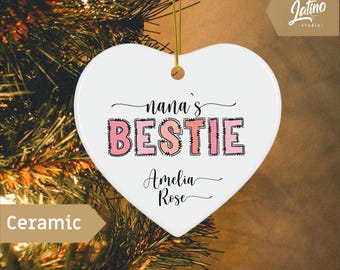 May include: A white ceramic heart-shaped ornament with the words "nana's BESTIE" in black and pink lettering. The name "Amelia Rose" is written below. A gold string is attached for hanging. The word "Ceramic" is also visible.