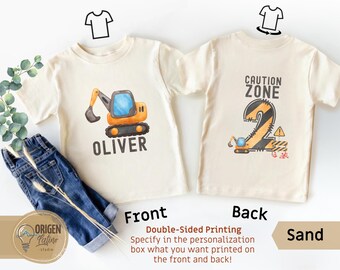 May include: Sand-colored toddler t-shirts with front and back designs. The front features an orange and blue excavator with the name "OLIVER". The back displays "CAUTION ZONE" with a number 2 and an excavator graphic. Includes the text "Double-Sided Printing".