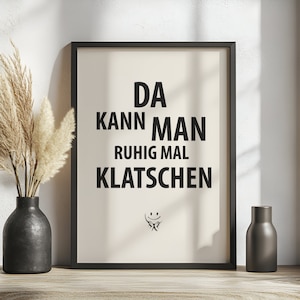 May include: Framed art print with the German phrase "DA KANN MAN RUHIG MAL KLATSCHEN" in bold black letters on a cream background. A small smiley face graphic is below the text. The print is in a black frame, with a black vase and decorative grass to the left.