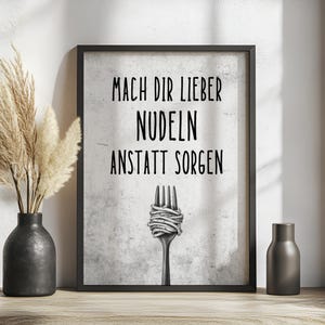 May include: Black framed print with the text "MACH DIR LIEBER NUDELN ANSTATT SORGEN" above a fork with spaghetti. A black vase with dried plants and a small black bottle are on a wooden surface.