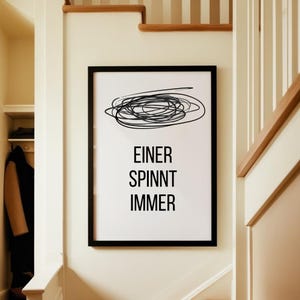 Könnte beinhalten: Ein gerahmter Kunstdruck mit schwarzem Rand, der an einer weißen Wand hängt. Der Druck zeigt ein schwarzes Gekritzel-Design über dem deutschen Text "EINER SPINNT IMMER" in einer fetten, serifenlosen Schriftart. Das Kunstwerk wird in einem Flur gezeigt.