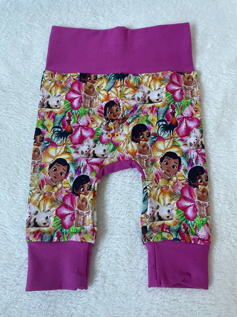 May include: Baby pants with a vibrant floral print featuring cartoon characters, flowers, and animals in shades of pink, yellow, and green. The waistband and cuffs are a solid magenta color.