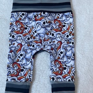 May include: White baby leggings with a black and gray striped waistband and cuffs. The leggings feature a repeating pattern of a cartoon ghost with a red tongue and a black eye.