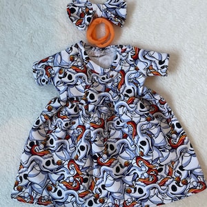May include: A white, blue, and orange patterned dress with a matching bow and orange hair tie. The dress features a print of a cartoon character. The dress has short sleeves and a flared skirt, suitable for a child.