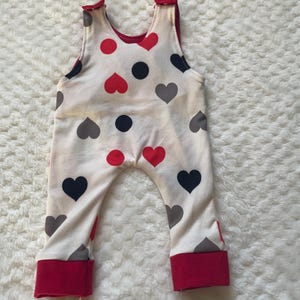 May include: White baby overalls with a red, black, and gray heart pattern. The overalls have red trim around the straps and leg openings.