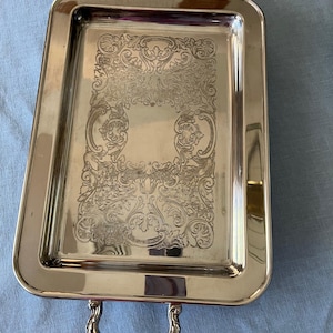 Puede incluir: Una bandeja de servir plateada con diseños grabados ornamentados y dos asas. La bandeja es rectangular y tiene un acabado pulido.