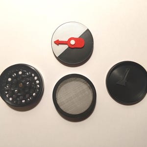 May include: A four-piece black herb grinder set. The set includes a top piece with a red arrow, a grinding chamber, a mesh screen, and a bottom piece with a scraper. The top piece has a black and white design.