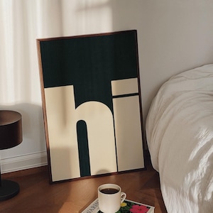 Op de afbeelding: Een ingelijst drukwerk met het woord "hi" in een vet, wit lettertype op een donkergroene achtergrond. De print ligt op een houten oppervlak naast een witte koffiemok en een boek met de titel "Warhol".