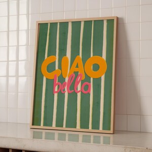 May include: A framed art print with a green and white striped background. The words "CIAO bella" are in orange and pink. The artwork is displayed on a white tiled wall.