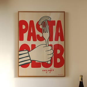 Op de afbeelding: Een keukeninterieur met een ingelijste poster met de tekst "Pasta Club". Een terracotta kom vol sinaasappelen staat op een marmeren aanrecht, met losse sinaasappelen verspreid. Twee wijnflessen zijn ook aanwezig.