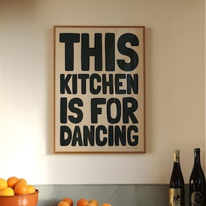 Op de afbeelding: Keukeninterieur met een ingelijst bord met de tekst "THIS KITCHEN IS FOR DANCING". Een terracotta kom gevuld met sinaasappels staat op een aanrecht, met losse sinaasappels verspreid. Er staan ook twee wijnflessen.