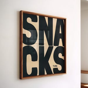 May include: A framed print with the word "SNACKS" in large, black, bold letters. The letters are stacked on top of each other. The background is a light beige colour. The frame is a dark wood colour. The text "TIME" is in smaller letters below the word "SNACKS".