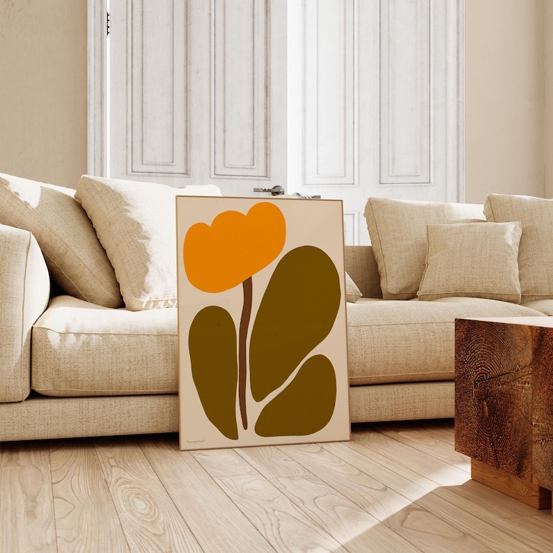 Peut inclure: Impression d'art abstrait repr&eacute;sentant une fleur stylis&eacute;e avec des p&eacute;tales orange et des feuilles vertes. L'impression est encadr&eacute;e et accroch&eacute;e &agrave; un mur dans un salon.